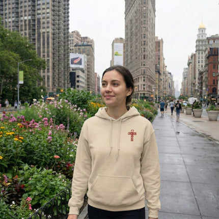Person wearing a beige hoodie with a red logo on a city street with buildings and flowers in the background